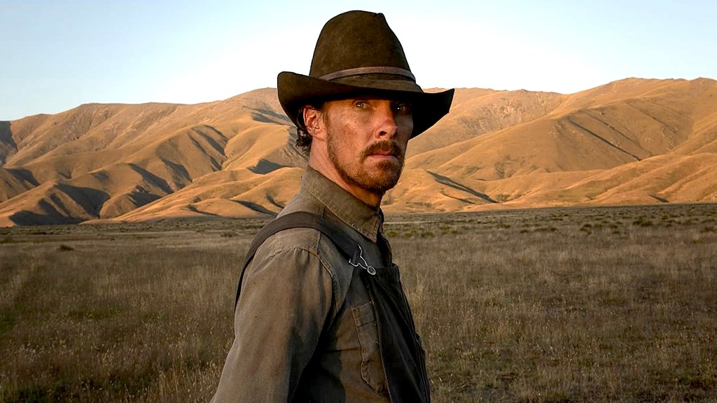 Benedict Cumberbatch as Phil Burbank wearing overalls and brown hat with green grass and brown mountains behind him